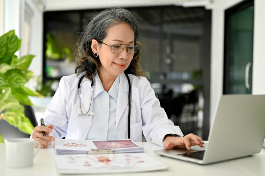 Professional Asian aged female doctor in uniform working in the doctor's office, using laptop computer to look at EHRs