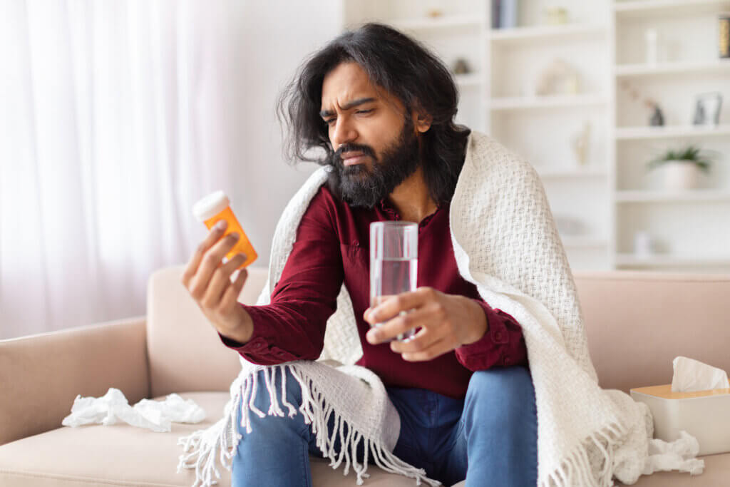 A nonadherent Indian man wrapped in a blanket looks confusedly at a bottle of pills while holding a glass of water, illustrating illness and treatment