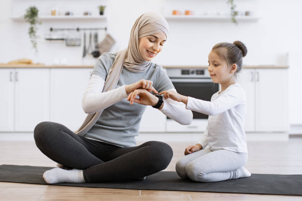 Muslim female with daughter checking workout achievements on sports watch while sitting on yoga mat in kitchen. Mother and her girl taking advantages of home training using fitness tracker and IoMT